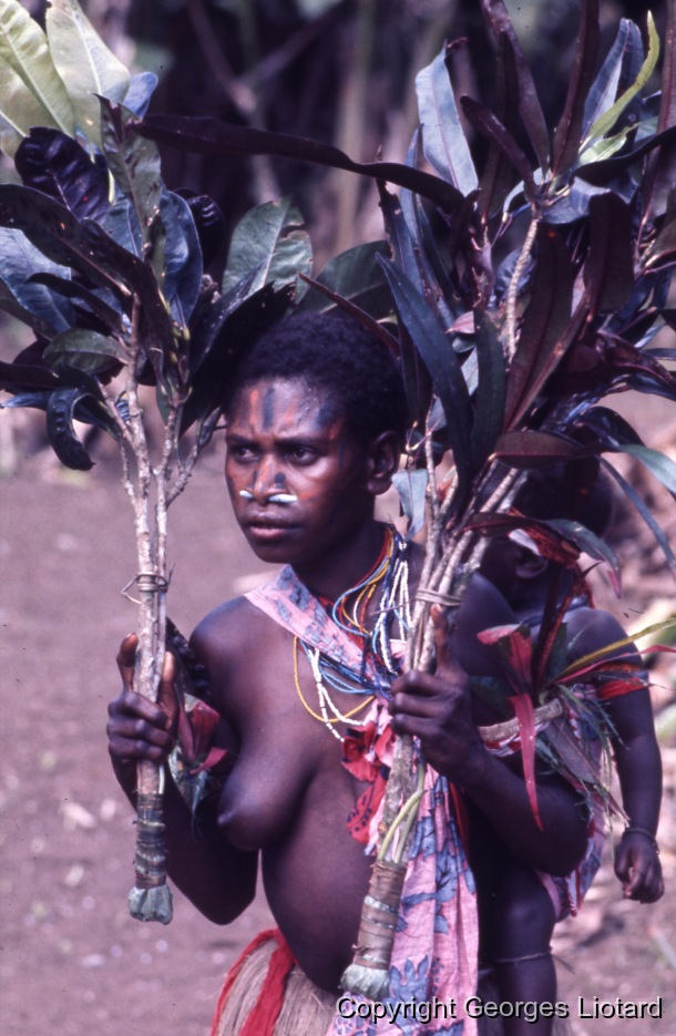 Funérailles à  Malakula (Malekula, Mallicolo) Vanuatu / Jeune femme: Jeune femme visage peint portant un enfant dans le dos et brandissant des rameaux de feuille de croton. / Liotard, Georges / Lamap, Malekula, Vanuatu
