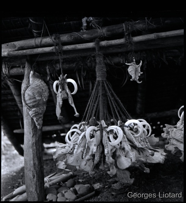 Ilôt  MALO / Le petit auvent, au toit duquel sont suspendus de nombreux pendants de mach (signe du haut grade obtenu). Y sont également suspendus des coquillages (conques et araignées de mer). Posés à même le sol, on voit des assomoirs en bois en forme de matraque / Georges Liotard / Villages d'Abunaress et de Narombe - Ile Malo - Vanuatu